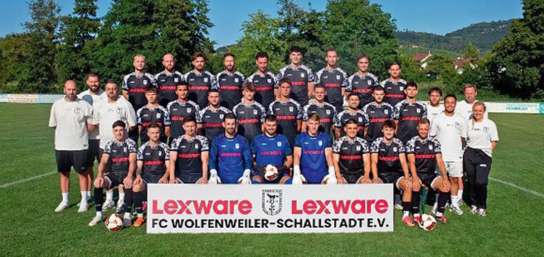 Eine Fu ballmannschaft mit Spielern in roten Trikots und Trainern in wei en Shirts posiert auf einem Rasenfeld hinter einem Schild mit der Aufschrift SC Hofstetten vor einer bewaldeten H gellandschaft. Beschreibung von KI generiert
