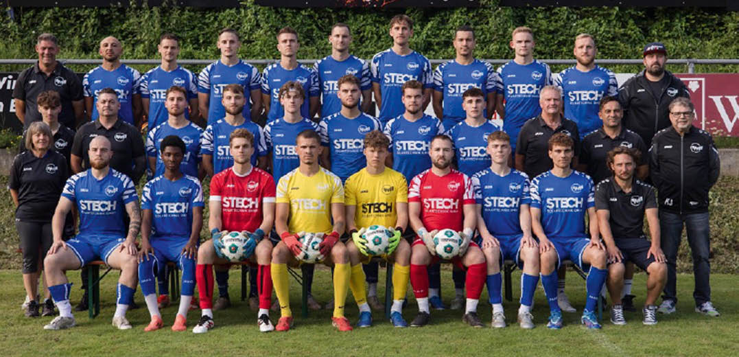 Eine Gruppe von Erwachsenen in blauen, gelben und roten Sporttrikots posiert in drei Reihen auf einem Fu ballfeld vor einem gr nen Hintergrund. Beschreibung von KI generiert